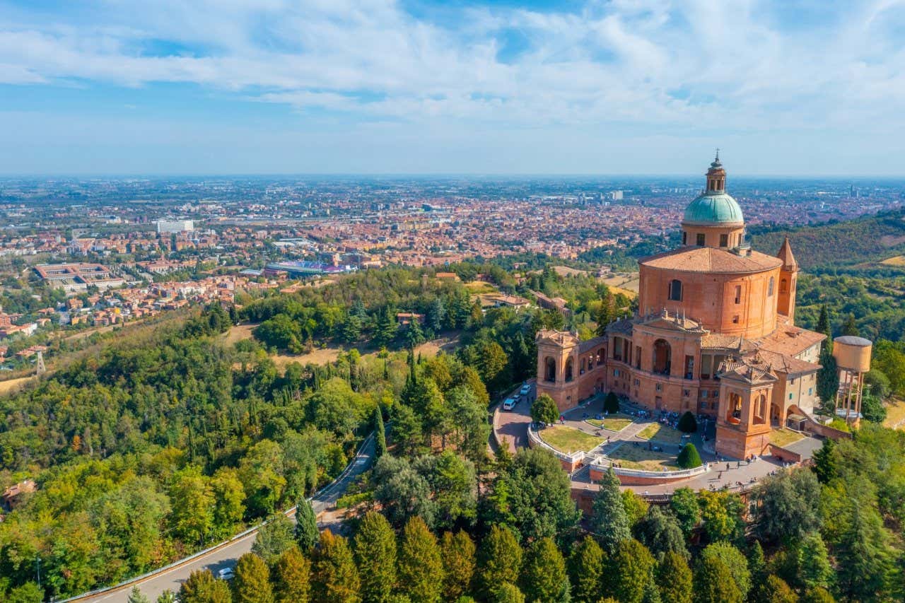 Veduta aerea del Santuario di San Luca a Bologna, circondato da una rigogliosa vegetazione verde e con la città sullo sfondo, sotto un cielo sereno e azzurro