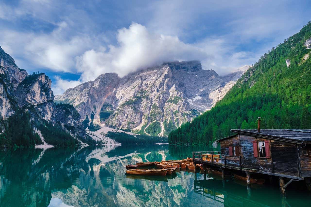 Un lago in alta quota con alcune barche in legno attraccate vicino a una palafitte e cime di montagne sullo sfondo coperte da nuvole