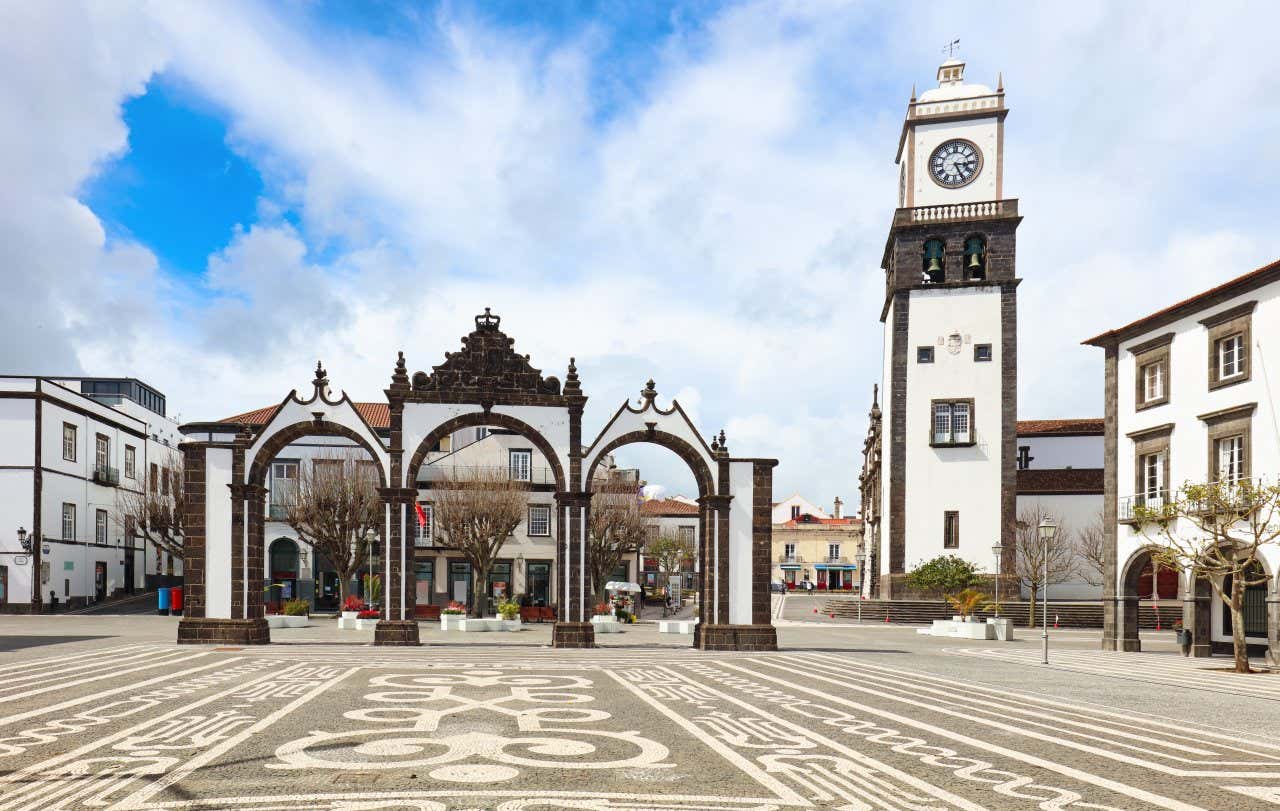 Piazza con pavimento decorato da motivi geometrici in bianco e nero, al centro della quale si erge un monumentale arco triplo in pietra scura. Sullo sfondo, si vede una torre dell'orologio bianca con dettagli in pietra scura. L'architettura circostante è in stile tradizionale, con edifici bianchi dalle cornici scure. Il cielo è parzialmente nuvoloso.