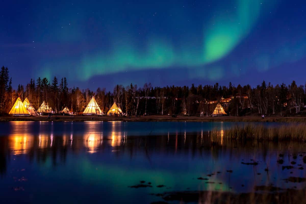 Um lago à noite, cercado por barracas em formato triangular e uma aurora boreal esverdeada no céu