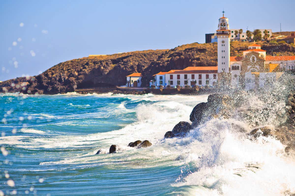 The waves of the sea breaking loudly on the coast next to the basilica of Candelaria, a church with a white façade.