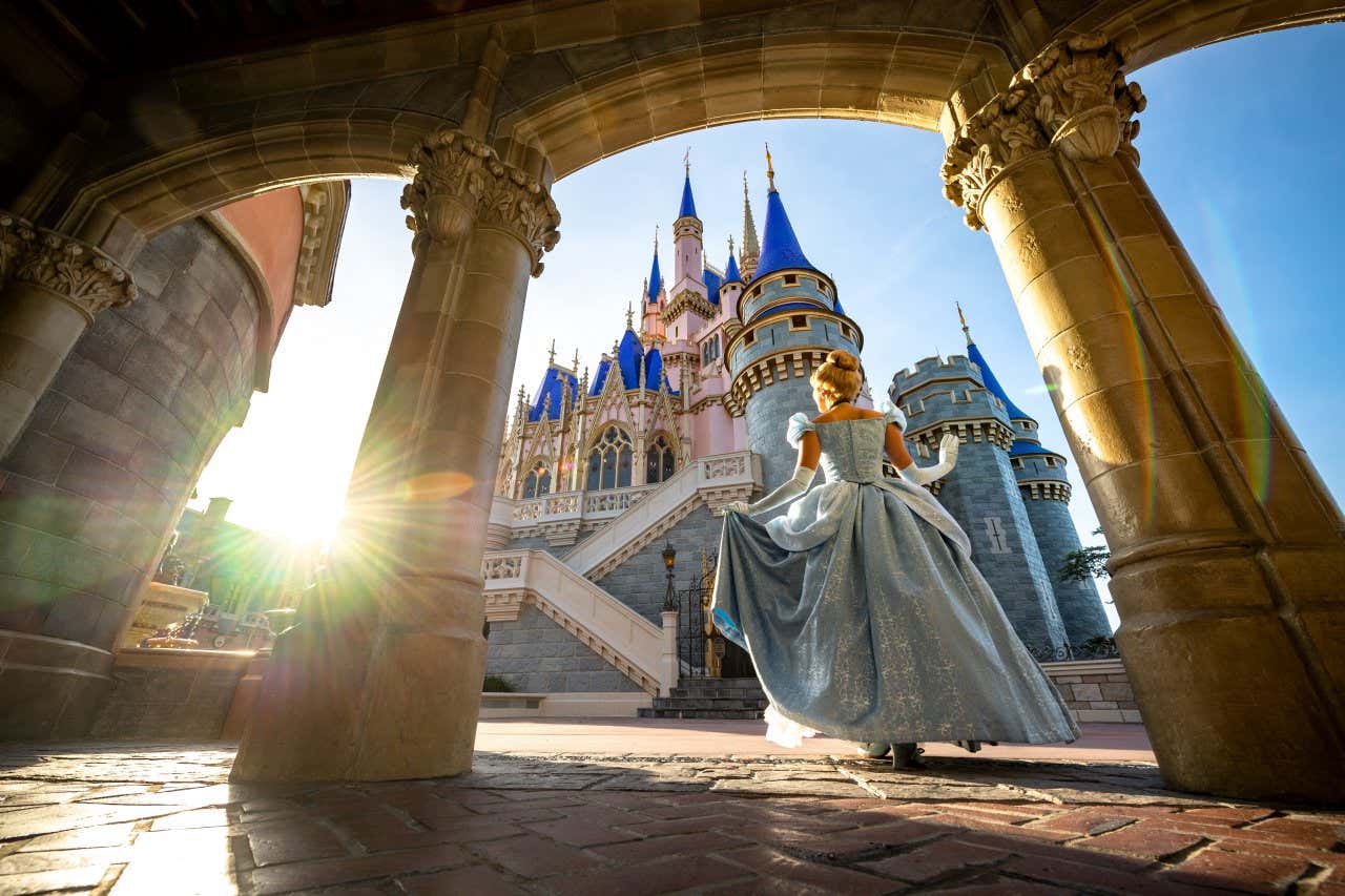 Una persona vestita da principessa, con un abito lungo e blu, cammina verso un castello fiabesco con torri blu e dorate, illuminata dalla luce del sole che tramonta, vista attraverso un arco di pietra