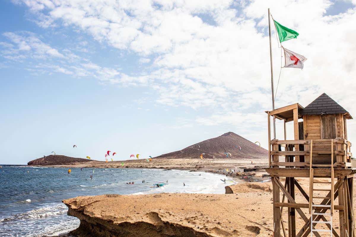 A lifeguard post, made of wood, next to a beach with several people kitesurfing and paragliding in El Médano.