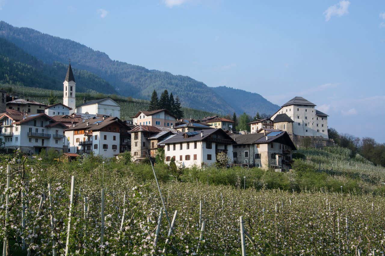 Vista di un piccolo villaggio di montagna circondato da campi coltivati, con case tipiche dai tetti spioventi e una chiesa con campanile. Sullo sfondo si vedono colline e montagne coperte di boschi, sotto un cielo sereno. A destra, un grande edificio storico, simile a un castello, domina la scena