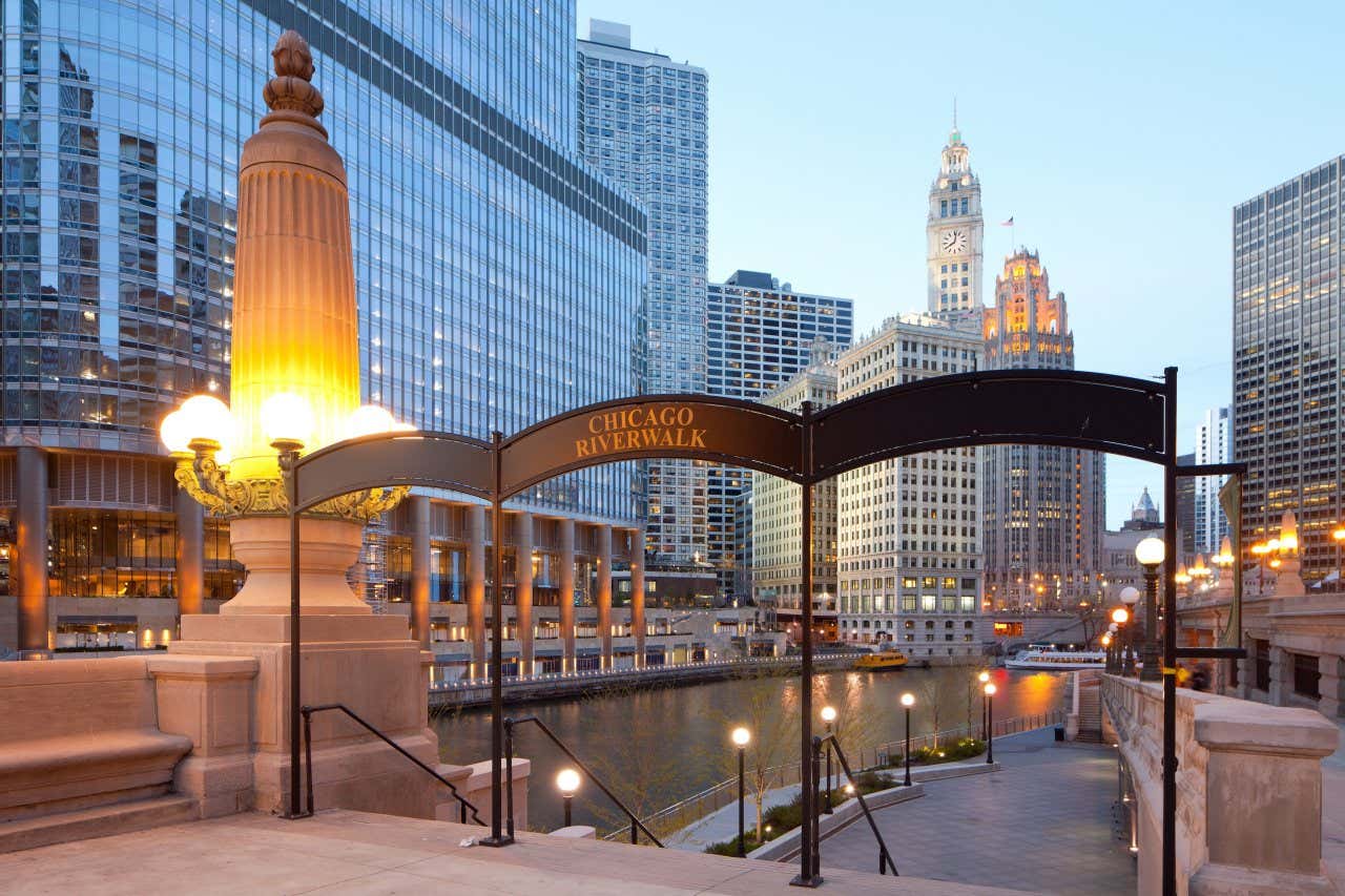 Placa da entrada do Chicago Riverwalk às margens do rio com os edifícios ao fundo ao entardecer