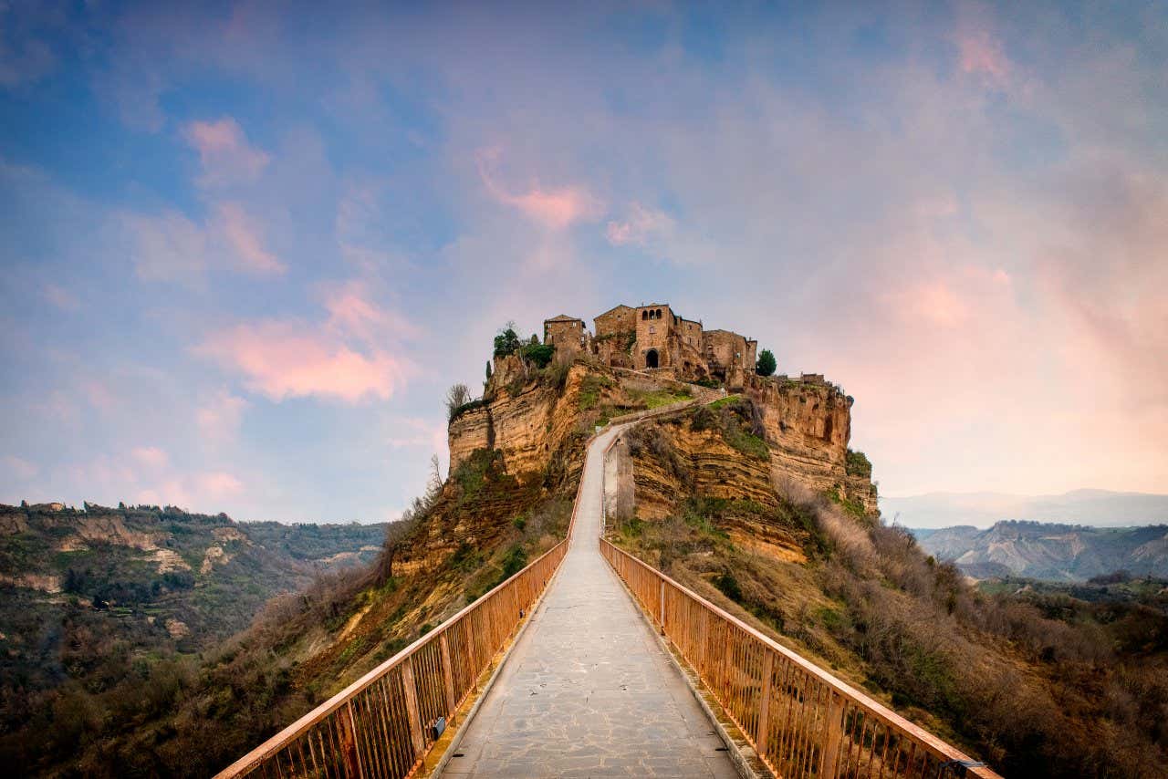 Primo piano prospettico di un ponte che conduce a un piccolo borgo in pietra arroccato su una stretta collina in tufo al tramonto