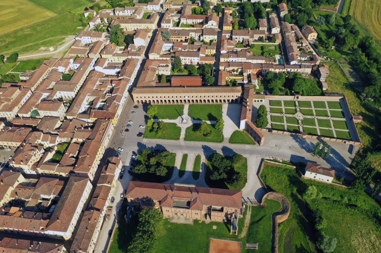 Vista aerea di una cittadina con edifici disposti in modo ordinato. Al centro dell'immagine si trova una grande piazza rettangolare con giardini curati e una statua centrale. Intorno alla piazza si vedono edifici storici con tetti in tegole rosse e cortili interni, e sul lato destro si nota una zona recintata con giardini geometrici