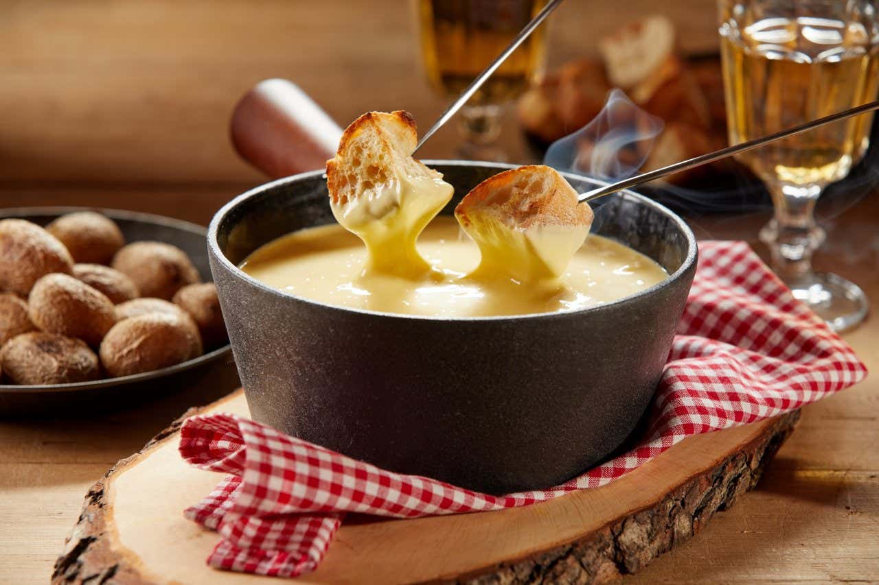 Uma panela com fondue de queijo em cima de um tecido xadrez vermelho e branco com dois garfos mergulhando o pão no fondue