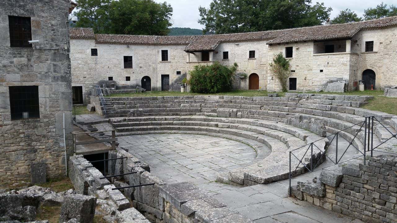 Scorcio di un anfiteatro romano in marmo grigio circondato da palazzi medievali in pietra dal tetto spiovente