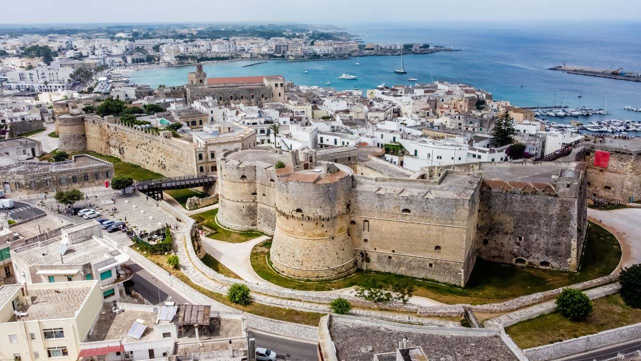 Vista dall'alto di una fortezza con masti e torrioni, immersa in un agglomerato urbano recente affacciato sul mare in un giorno con una foschia leggera