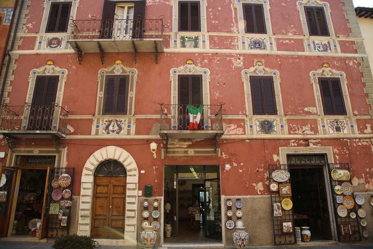 Facciata di un palazzo signorile di Deruta di colore rosso decorato con maioliche e affreschi. Al piano terra tre botteghe che espongono piatti di ceramica colorati