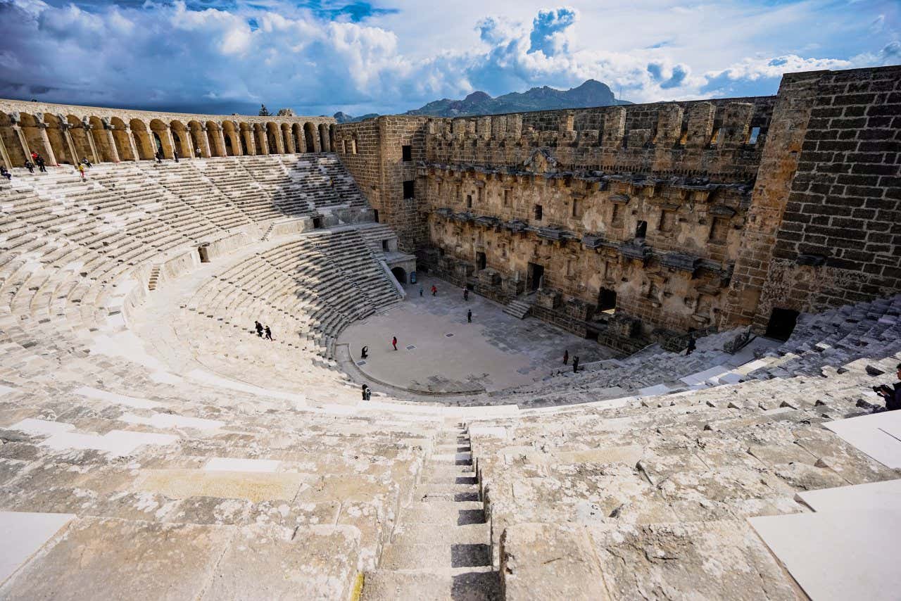 The Aspendos Theatre as seen from one of the top steps, with tourists walking within and a view of the landscape in the background.