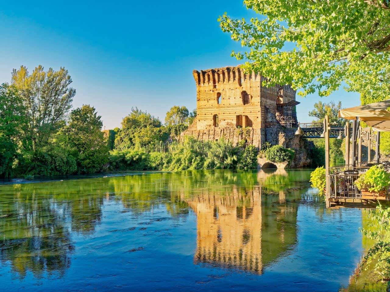 I resti di una mastio in pietra immerso nel verde sul lato di un fiume con un pontile in ferro che lo connette alla sponda opposta. Sull'acqua del fiume c'è il riflesso dell'antico edificio illuminato dalla luce del tramonto