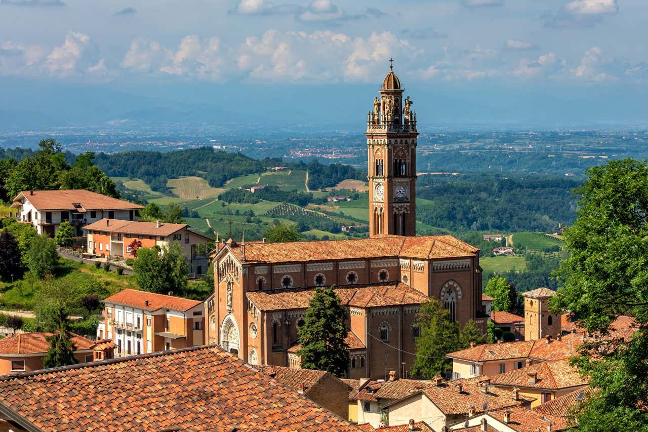 Vista panoramica di una città collinare con una grande chiesa in mattoni rossi al centro, caratterizzata da un campanile alto con orologio. Intorno si trovano case dai tetti in tegole rosse e, sullo sfondo, un paesaggio verdeggiante di campi e colline