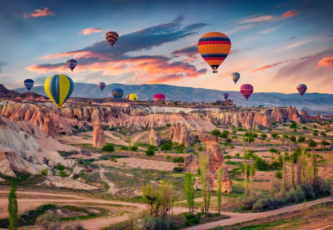 An expansive landscape in Cappadocia, with hot air balloons all over, and a cloudy evening sky in the background.