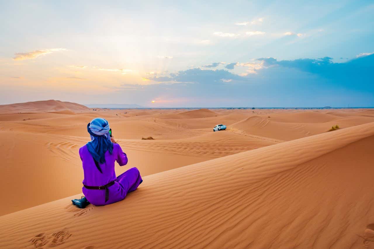 Una persona di spalle e con un turbante sulla testa si gode il tramonto sul deserto seduto a gambe incrociate su una duna di sabbia di un'intenso colore arancione