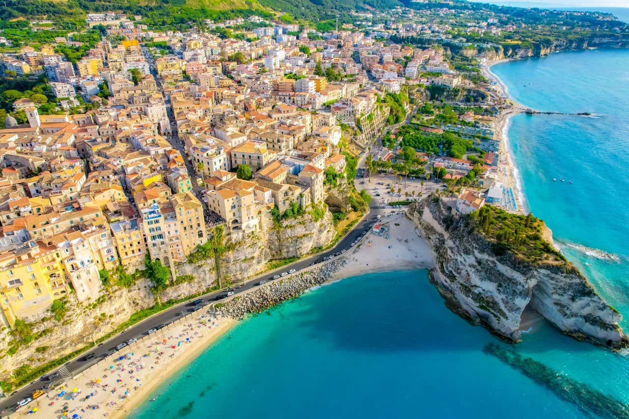 Vista dall'alto di una città affacciata su un mare cristallino e una costa con spiagge di sabbia e scogli, con alcune formazioni rocciose che interrompono la lingua di sabbia