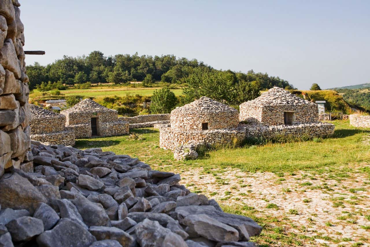 Vista di piccole capanne costruite con pietre bianche connesse da bassi muretti fatti con le medesime pietre all'interno di una piccolo spiazzo immerso nel verde