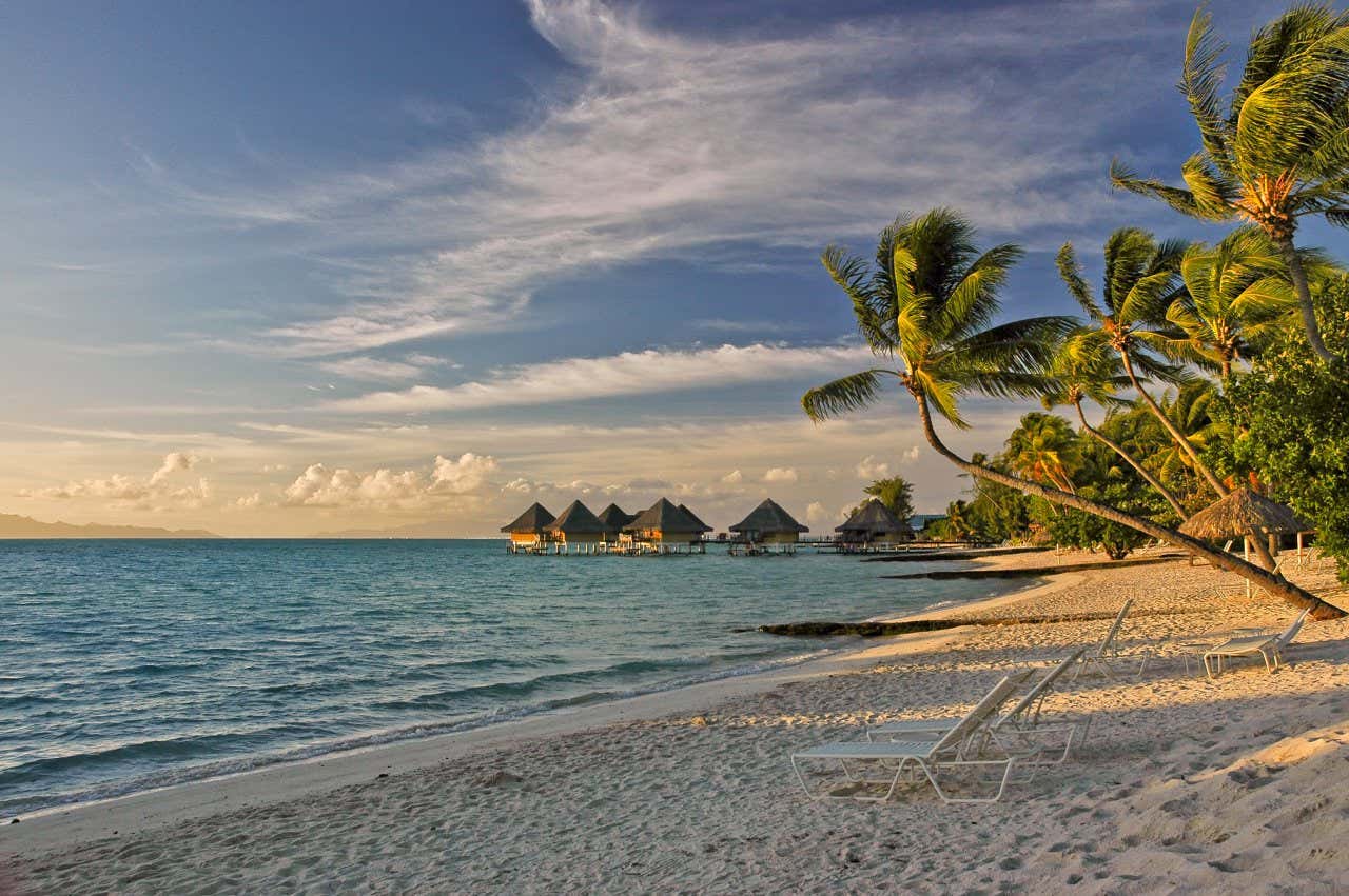 Palmiers et transats sur la plage de Matira, l'une des plus belles de France, au coucher du soleil, maisons sur îlots en arrière-plan