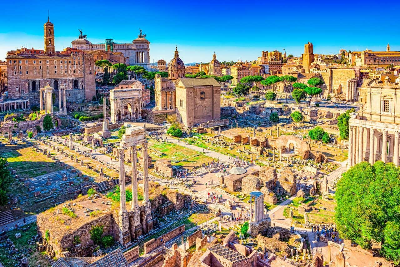 An aerial view of the Roman Forum, full of ruins of pillars and buildings, under a blue sky.