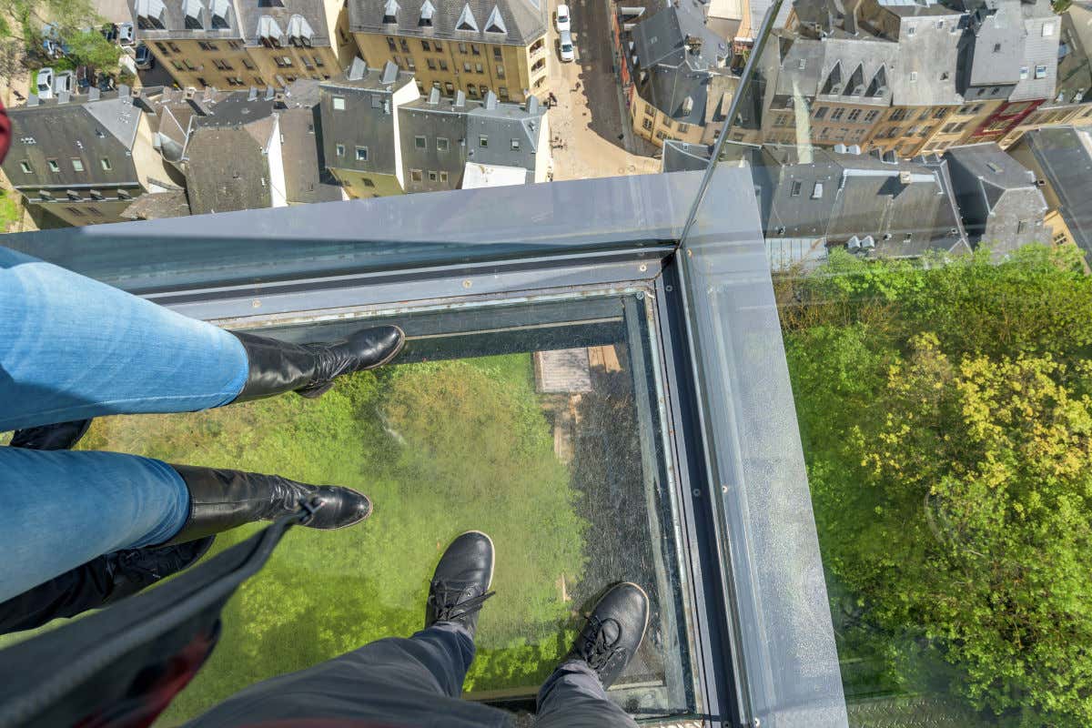 Quatro pernas apoiadas no piso de vidro de um elevador panorâmico
