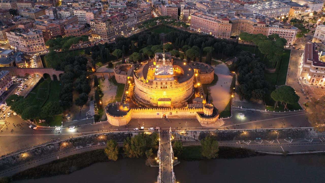 An aerial view of Castel Sant'Angelo lit up at night time, surrounded by a park of tall trees and vegetation.