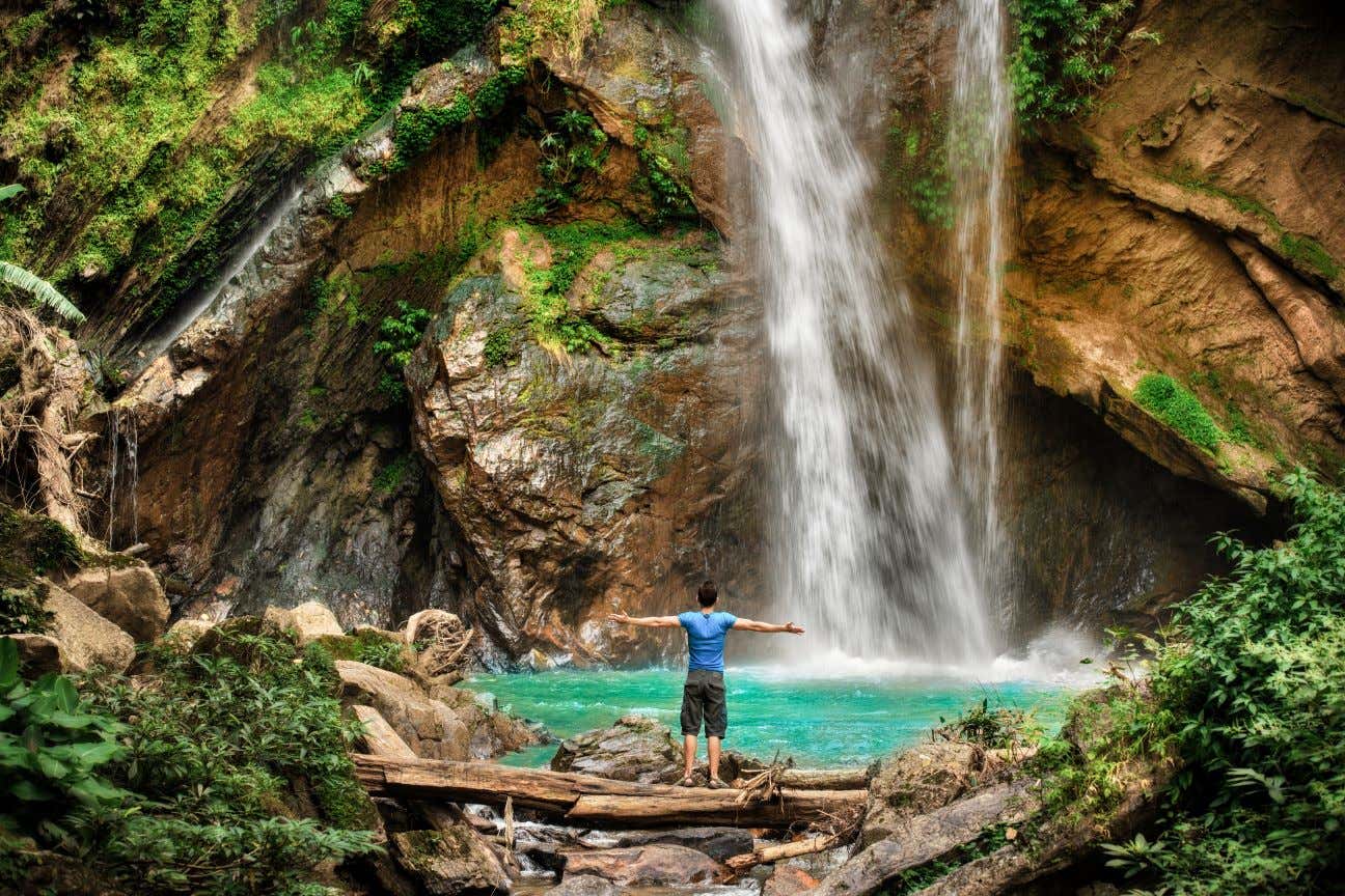 Un hombre de espalda expande sus brazos frente a una cascada y una laguna de aguas turquesas