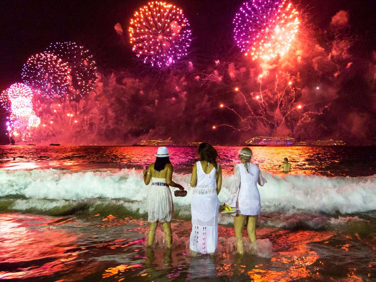 In primo piano tre donne di spalle e vestite di bianco camminano dentro l'acqua del mare ammirando in lontananza i fuochi d'artificio color fucsia