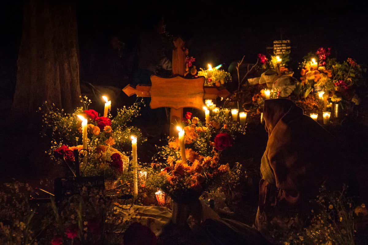 Uma mulher com um cobertor comemorando o Dia dos Mortos em um cemitério decorado com flores e velas.