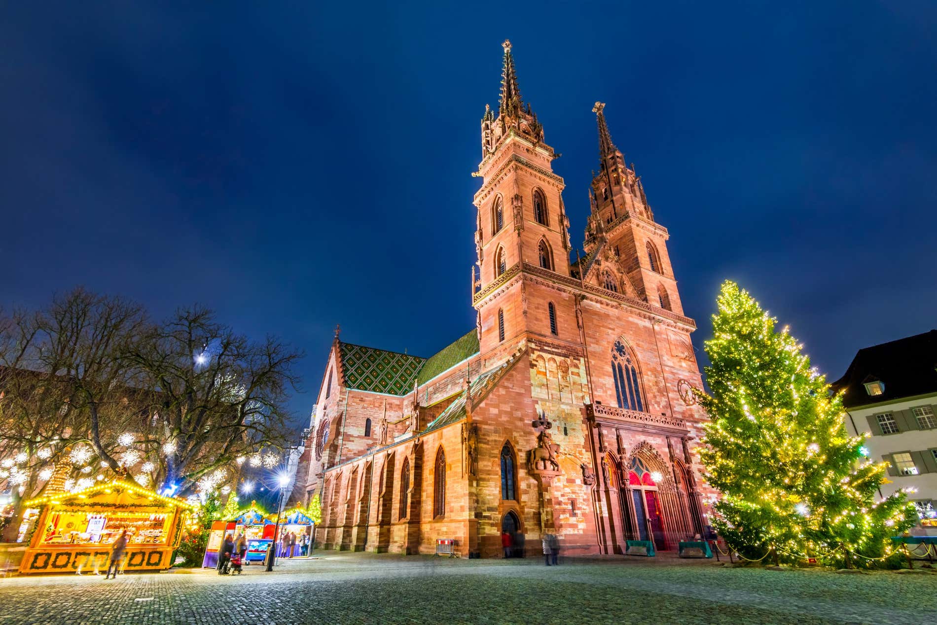 Una piazza del centro di Basilea fotografata dal basso. Al centro una cattedrale gotica con due campanili e davanti un grosso abete ricoperto di luci, sul lato sinistro invece delle piccole bancarelle decorate con luminarie e alcune persone che vi passeggiano davanti