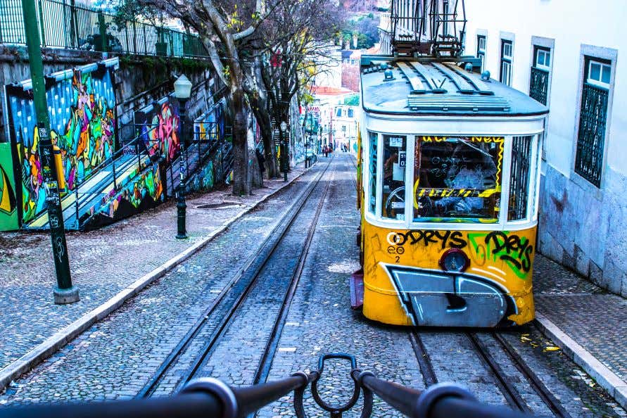 A view of a steep hill in Lisbon city with street art on the walls and graffiti on an old yellow tram.