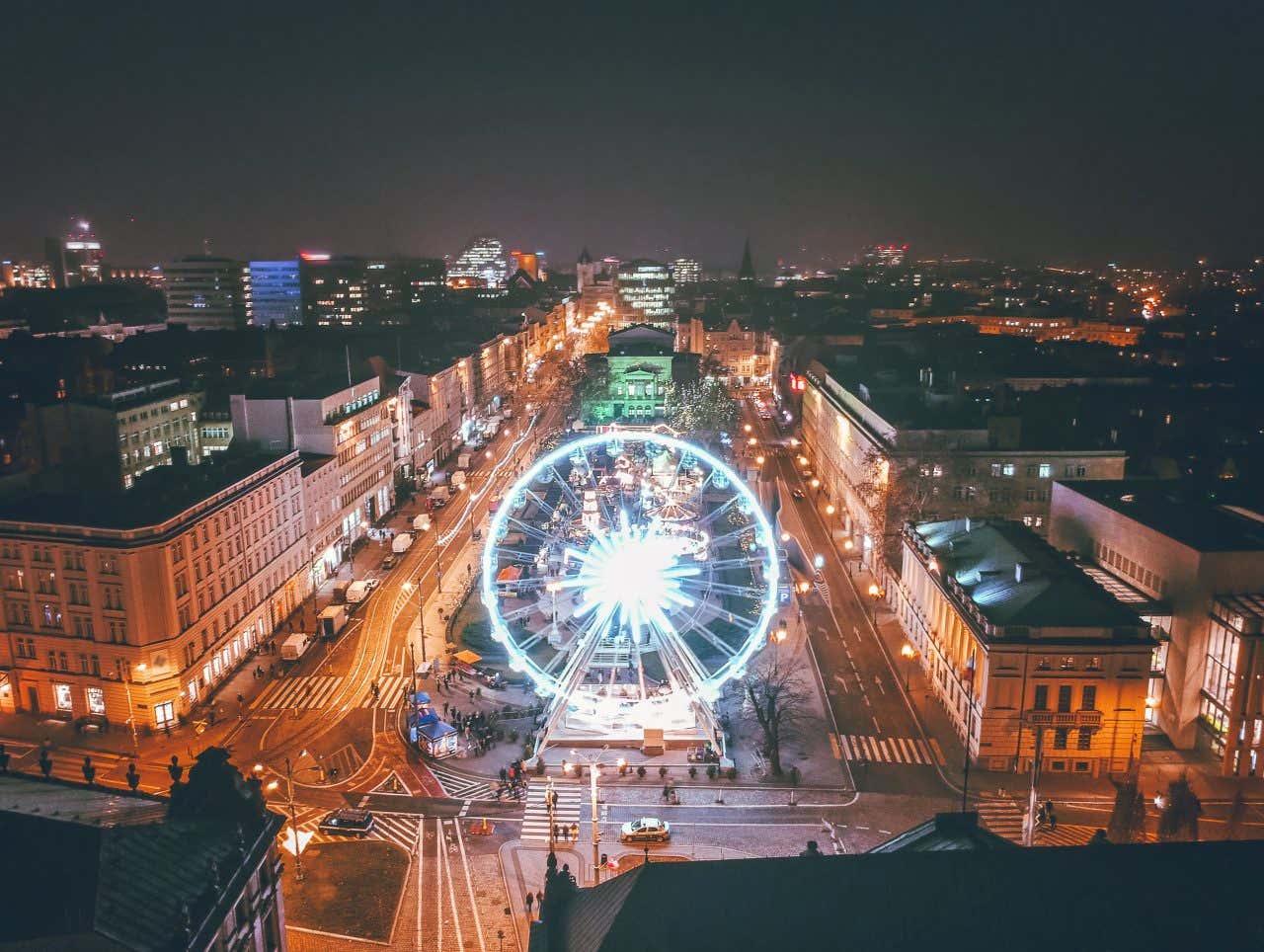 Foto notturna di una piazza gremita di bancarelle di legno, luminarie, alberi decorati e una ruota panoramica sullo sfondo