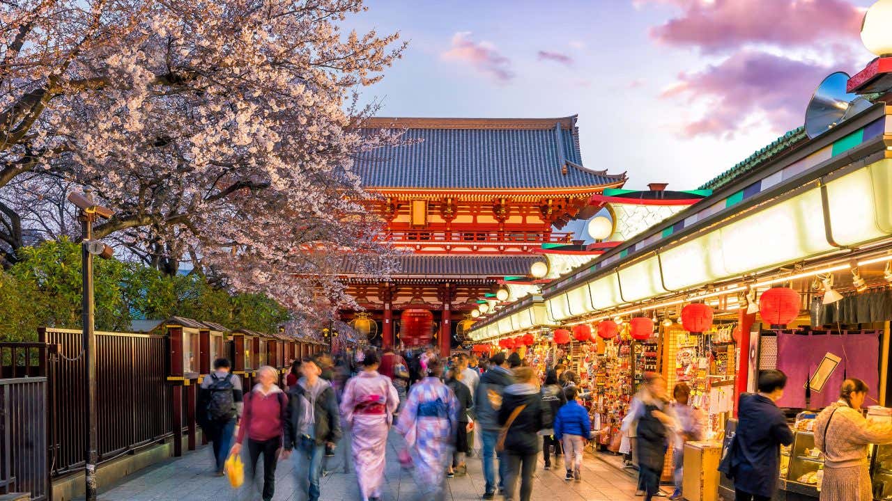 Una strada costeggiata da bancarelle decorate con lanterne rosse e percorsa da persone in abiti tradizionali e turisti occidentali. In fondo di vede un tempio a forma di pagoda rosso