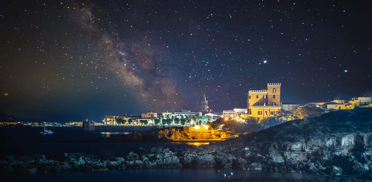 La città di Alghero di notte, affacciata sul mare e illuminata dal cielo stellato