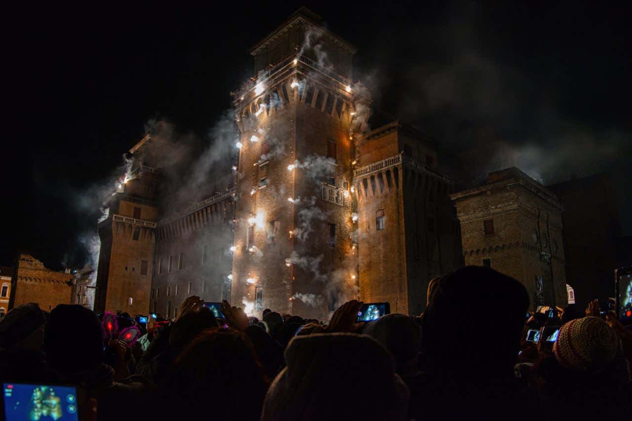 Il torrione di una rocca medievali coperto di sbuffi di fumo e piccole fiammelle, illumina la notte mentre una folla di persone, in basso, riprende la scena con il telefono