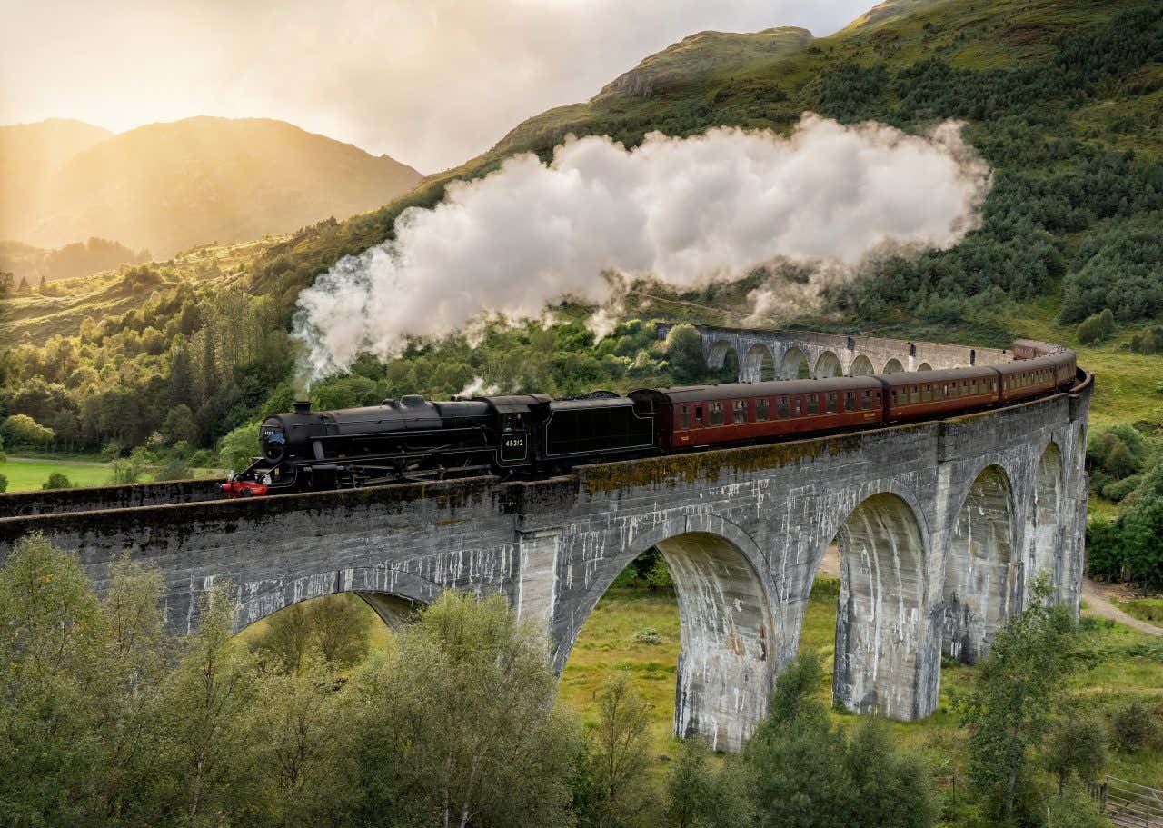 A view of a steam train on a stone bridge going through a hilly landscape, along the West Highland Train line, one of the best train journeys in Europe