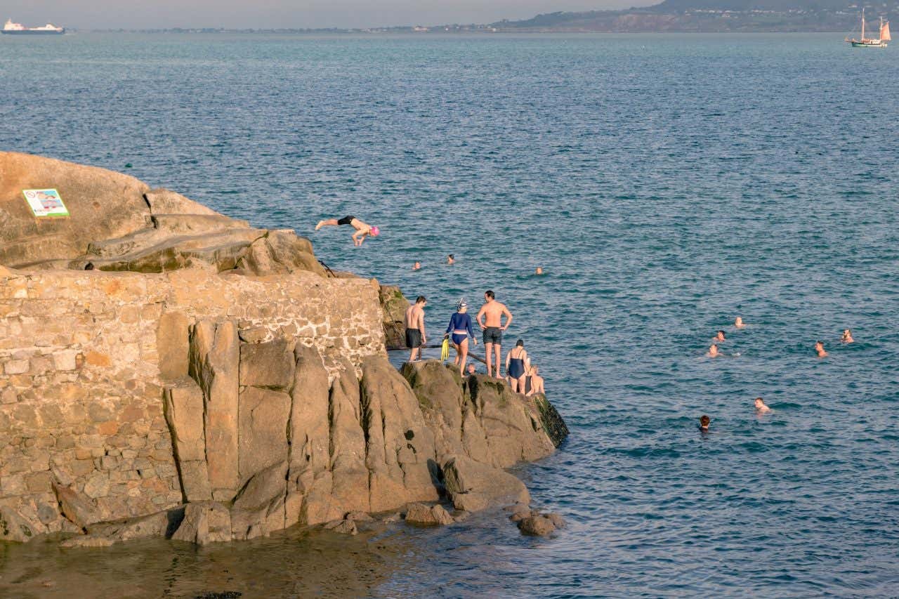 Persone che si tuffano da uno sperone roccioso mentre altre si fanno il bagno in mare