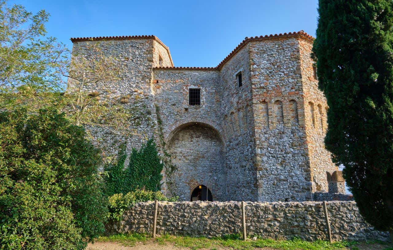 Le mura esterne di un antico castello medievale con ai lati due torri in pietra e un basso muro di cinta. Attorno alberi, edera e cespugli