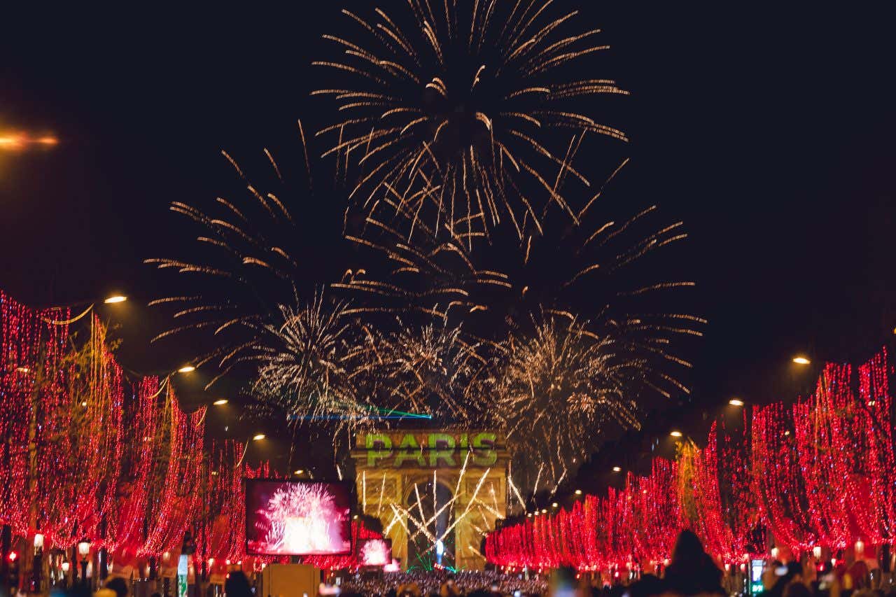 Vista degli Champs Elysées illuminati con luci rosse, mentre sullo sfondo c'è un arco di trionfo romano con la scritta "Paris" proiettata sopra e i fuochi d'artificio sullo sfondo