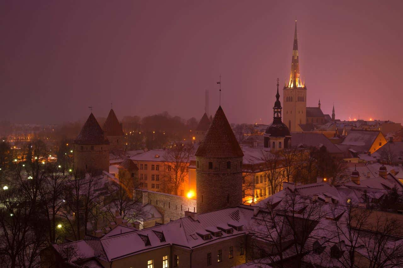 I tetti e le torri medievali del centro di Tallinn ricoperti di neve e fievolmente illuminati da luci di lampioni. Sono immersi nella nebbia in un'atmosfera invernale e romantica