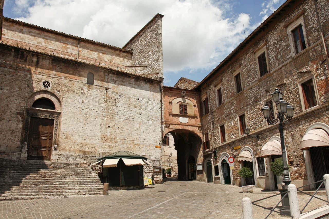 Una piazza in una cittadina storica, circondata da edifici in pietra. A sinistra c'è una grande chiesa con una facciata di pietra chiara e una porta di legno situata in cima a una breve scalinata. Al centro, si apre un arco che conduce a una strada interna, sormontato da un edificio con una finestra ad arco. Sulla destra, ci sono edifici in pietra con finestre rettangolari, tende sopra le porte e un lampione antico in primo piano. Il cielo è sereno con qualche nuvola bianca.