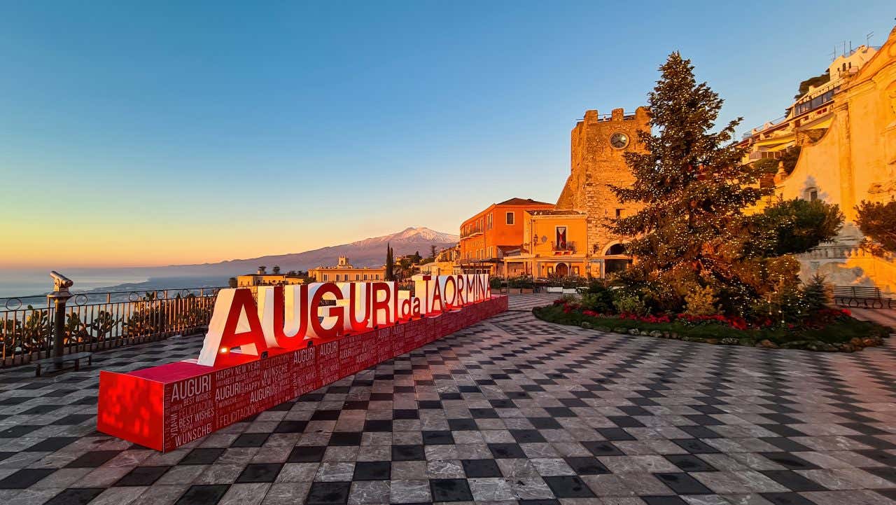 Un belvedere nel centro di Taormina con una grossa scritta rossa "Auguri da Taormina" e un albero di natale decorato al centro di una piccola porzione di giardino. Sullo sfondo una montagna illuminata dalle prime luci dell'alba