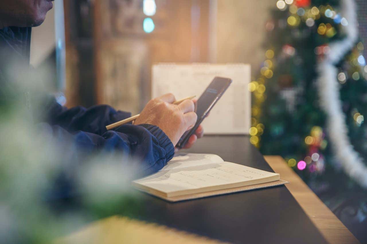 Un uomo cerca delle informazioni sul proprio smartphone mentre prende appunti su un'agenda all'interno di una stanza decorata con un albero di Natale