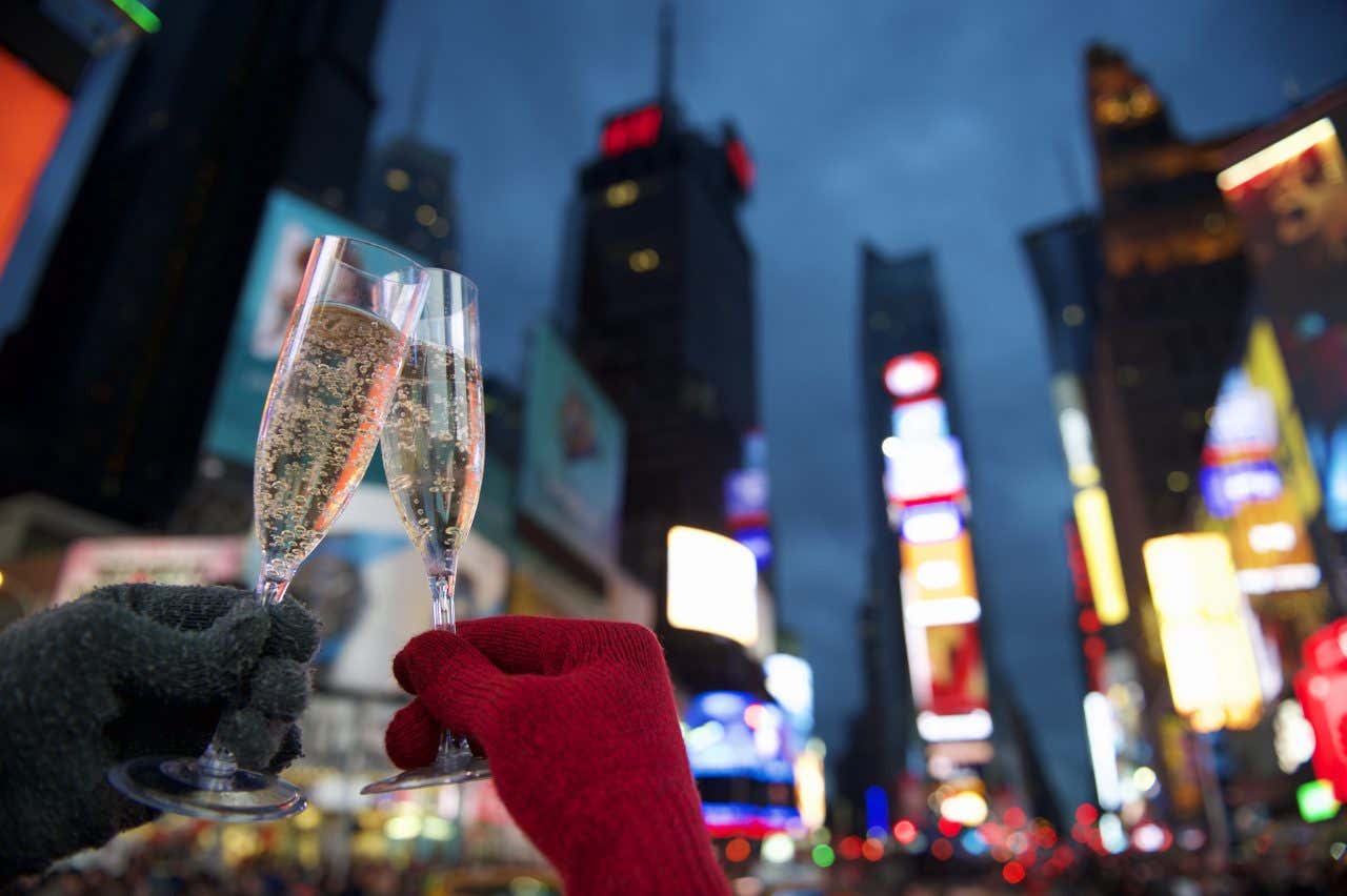 Due mani che indossano guanti di lana, uno grigio e l'altro rosso, reggono in mano due calici di champagne nell'atto di fare un brindisi. Sullo sfondo le luci dei grattacieli di Times Square, a New York