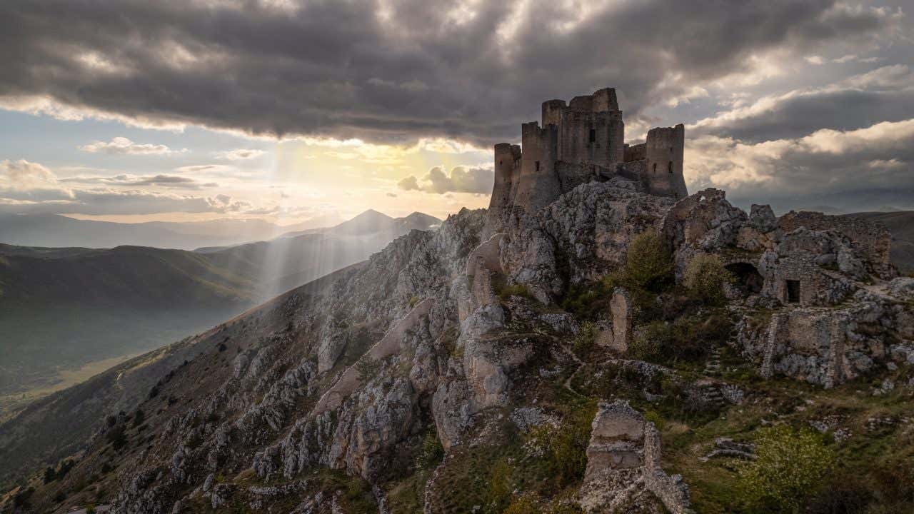 I resti di un antico castello con torri e mura di cinta, poggiati sulla cima rocciosa di un monte. Alle spalle un cielo nuvoloso trafitto da alcuni raggi di sole