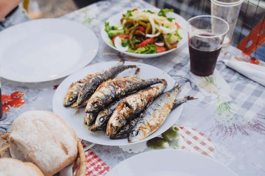 A plate of grilled sardines on a plate next to a plastic cup of wine and a salad and bread.