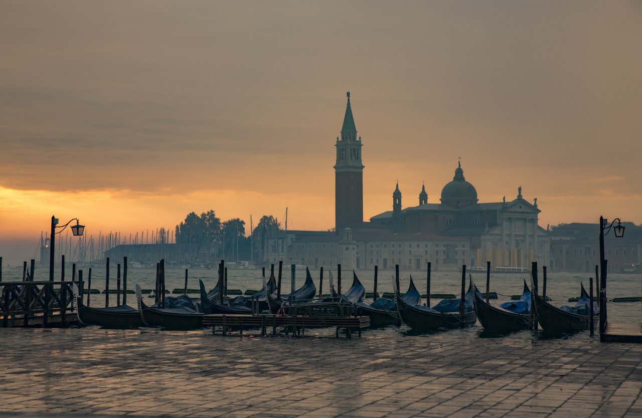 Gondole attraccate su un molo di Venezia e sullo sfondo il profilo di piazza San Marco offuscato dalla nebbia in una giornata nuvolosa con poca luce arancione che filtra dalle nubi sul lato sinistro dell'immagine