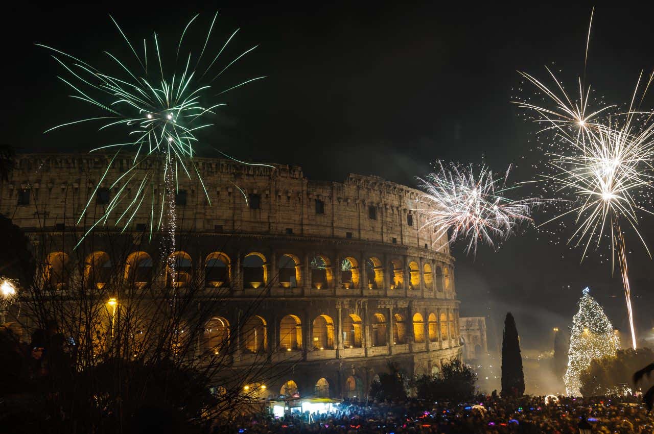 Il Colosseo di notte illuminati dai fuochi d'artificio. In basso una folla di persone in festa e un grosso albero di natale si intravede sullo sfondo