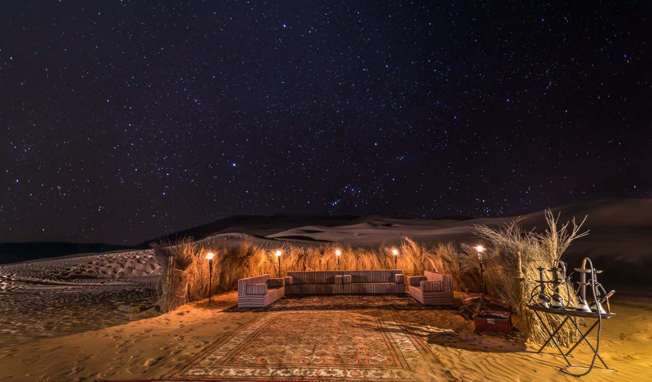 Uno spazio con divani e tappeti allestito in mezzo al deserto di notte. È illuminato da torce e nel cielo si vede un manto di stelle