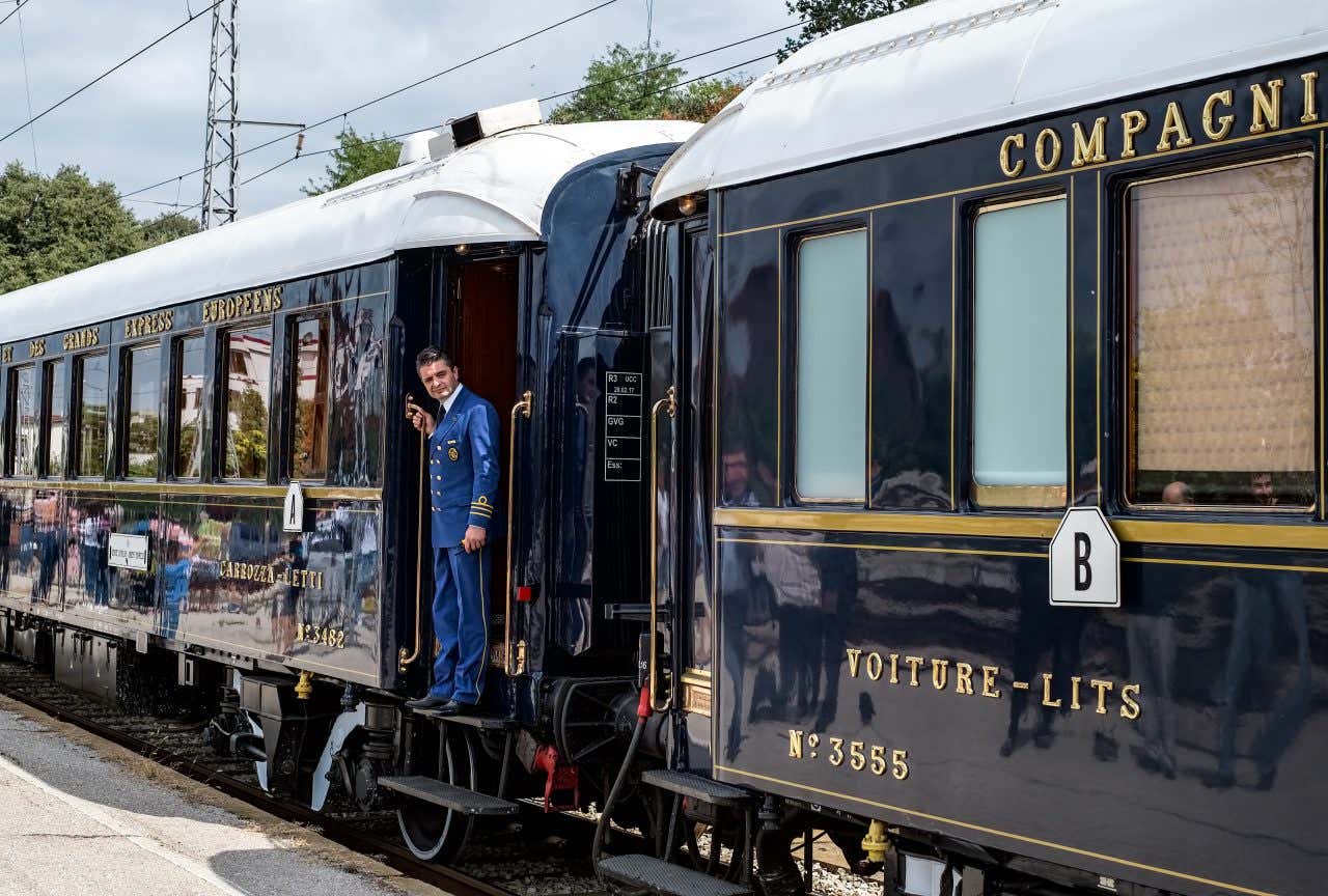 Two train carriages of the Orient Express, a train conductor in a blue uniform standing on the stairs waiting for guests to board the train